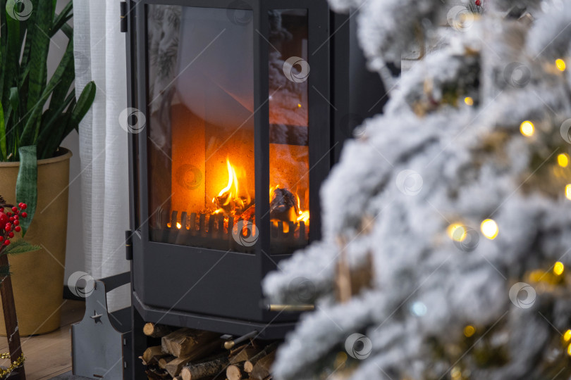 Скачать Металлическая черная печь, горящий очаг камин в белом цвете Праздничный интерьер дома украшен к Рождеству и Новому году, рождественская елка. дрова в поленнице, уют и отопление дома фотосток Ozero