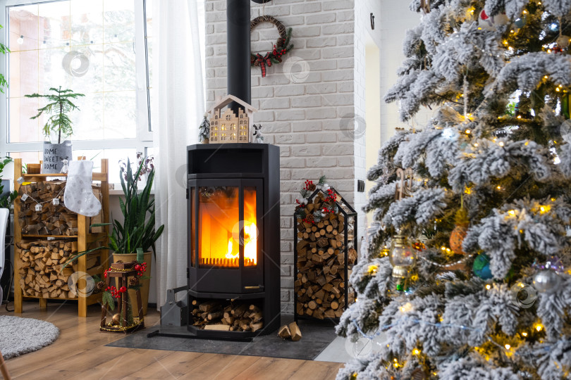 Скачать Металлическая черная печь, горящий очаг камин в белом цвете Праздничный интерьер дома украшен к Рождеству и Новому году, рождественская елка. дрова в поленнице, уют и отопление дома фотосток Ozero