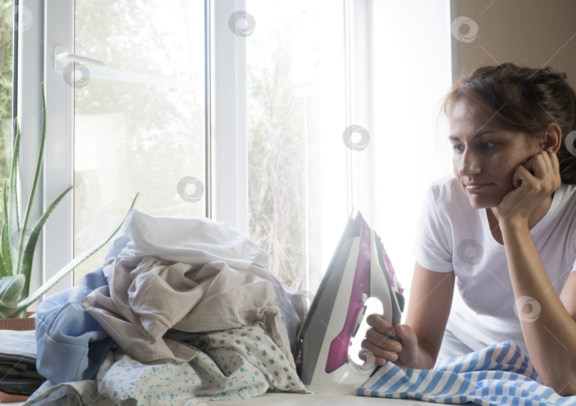 Скачать Разочарованная, усталая молодая женщина пристально смотрит на утюг и груду выглаженной одежды дома. Работа по дому. фотосток Ozero