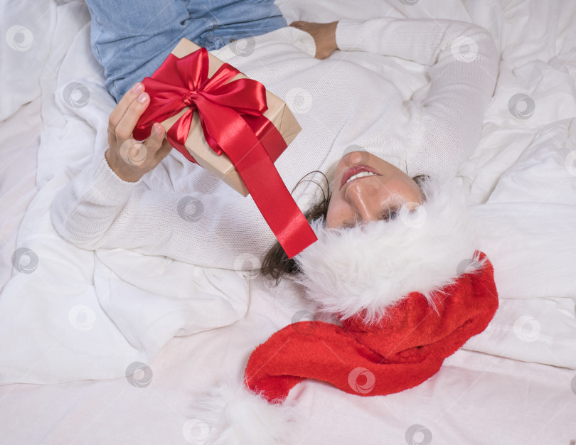 Скачать Красивая брюнетка в шапке Санта-Клауса радуется подарку в подарочной коробке с красной лентой на белой кровати, окруженной рождественскими украшениями. Концепция рождественских праздников. фотосток Ozero