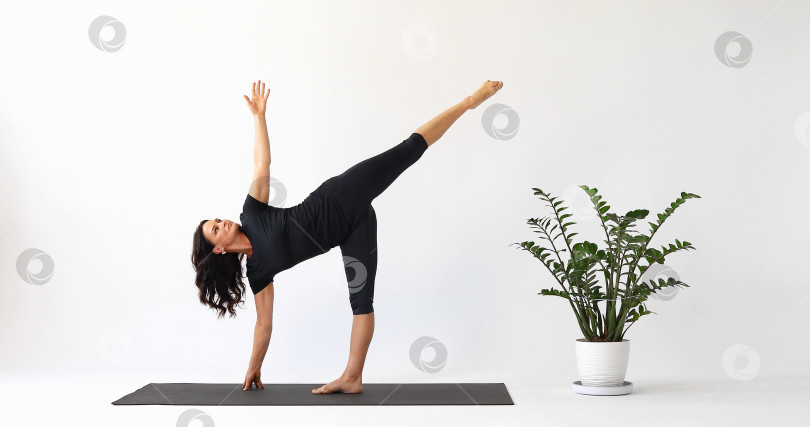 Скачать Инструктор по йоге выполняет упражнения Ардха Чандрасана, поза полумесяца, тренируется в коротких леггинсах и футболке, стоя на коврике в студии фотосток Ozero