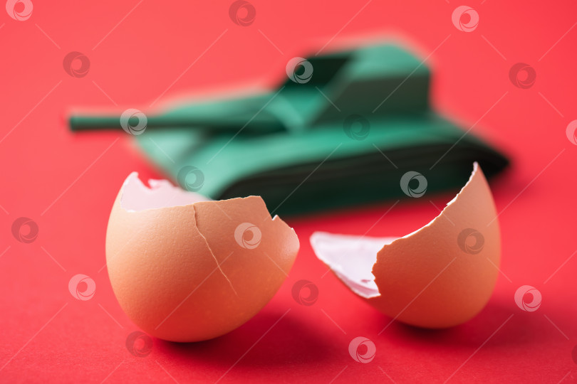 Скачать Разбитое пополам яйцо и игрушечный бумажный резервуар на красном фоне, концепт на тему хрупкого мира фотосток Ozero