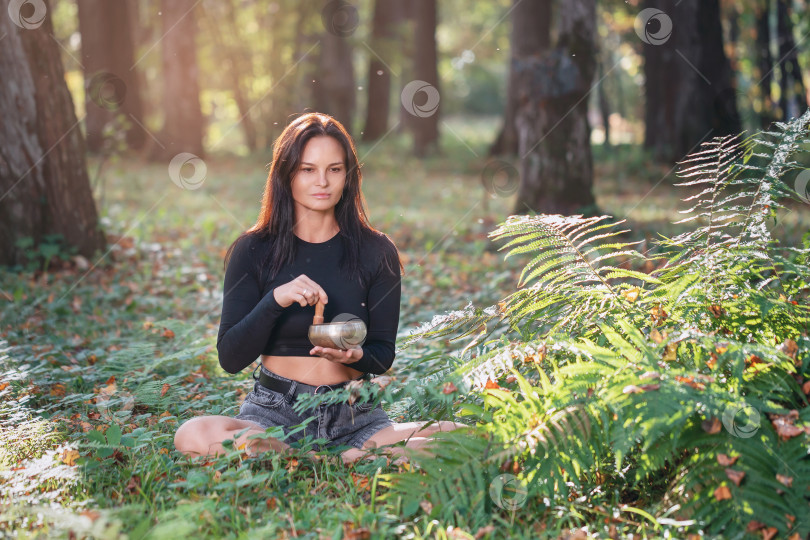 Скачать Женщина занимается звуковой медитацией с тибетской чашей, снимает стресс и улучшает общее состояние организма, занимается йогой в парке солнечным теплым осенним днем, сидя на поляне фотосток Ozero