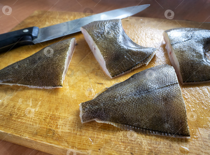 Скачать На деревянной доске лежат крупные куски камбалы и нож. Сырую рыбу нарезают ломтиками и обрабатывают для употребления в пищу. Приготовление рыбных блюд из камбалы. фотосток Ozero