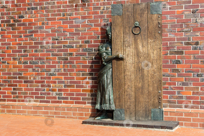 Скачать Бронзовая скульптура девушки, выглядывающей из-за двери в кирпичной стене. Скульптурная композиция "Тайное свидание". Туристическая зона "Рыбная деревня" на берегу реки Преголя. фотосток Ozero