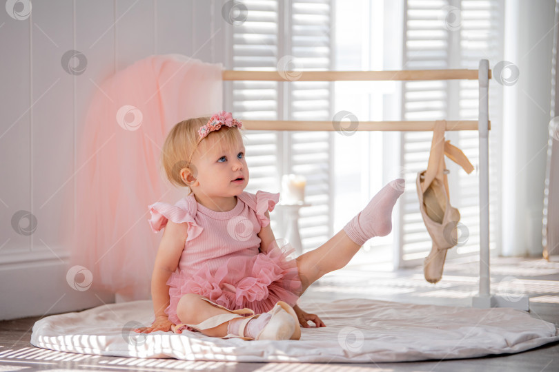 Скачать Симпатичная маленькая балерина в розовом балетном костюме сидит у станка в комнате и пытается надеть пуанты. Ребенок и балетный танец. Copyspace. фотосток Ozero