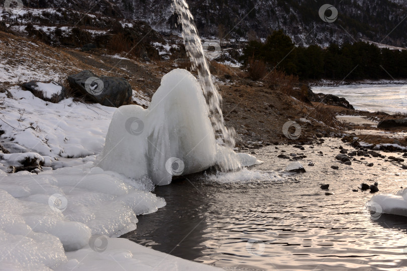 Скачать Поток воды небольшим ручейком стекает на ледяную глыбу на берегу красивой горной реки в ясный зимний день. фотосток Ozero