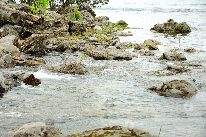 Скачать Камни и стволы поваленных деревьев на берегу большого озера с водоворотом, окруженного горами. фотосток Ozero