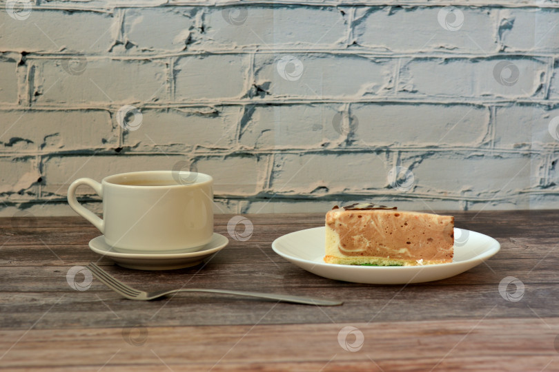 Скачать Чашка кофе на блюдце, вилка и тарелка с кусочком бельгийского крем-чизкейка на деревянном столе у кирпичной стены. фотосток Ozero