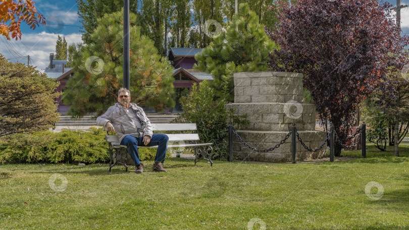 Скачать Взрослый мужчина в пуховике расслабляется, сидя на скамейке в парке. фотосток Ozero