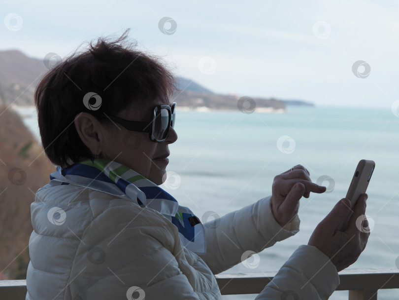Скачать Портрет в профиль. Пожилая женщина пользуется мобильным телефоном, делает селфи с балкона санатория на фоне моря. фотосток Ozero