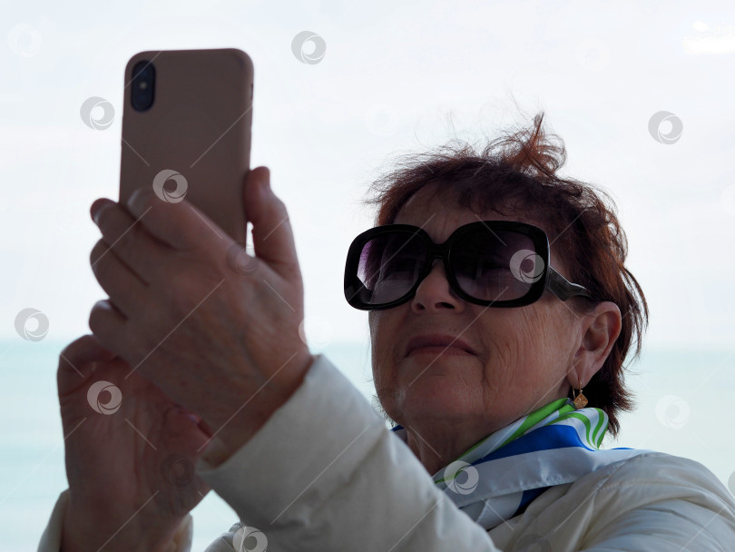 Скачать Пожилая женщина пользуется сотовым телефоном, делает селфи с балкона санатория на фоне моря. фотосток Ozero