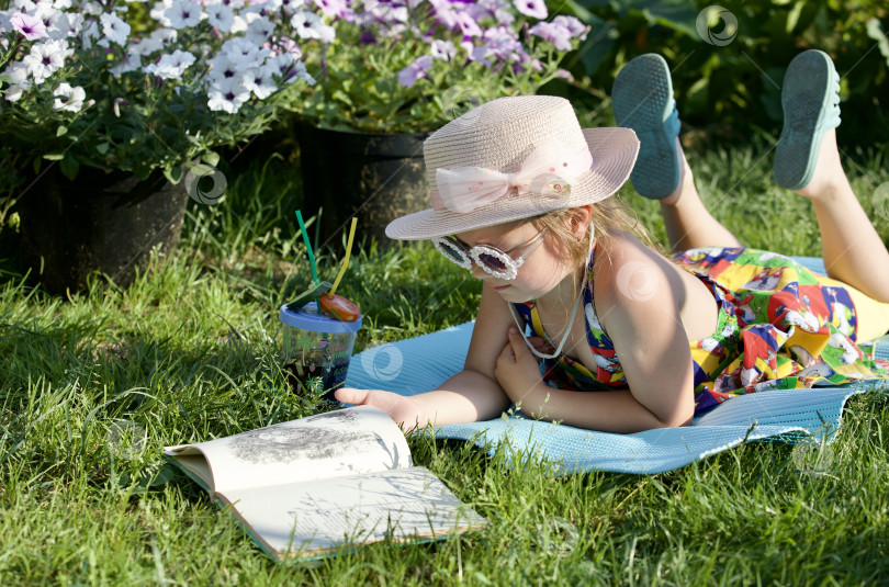 Скачать Девочка в ярком платье и шляпе читает книгу лёжа на траве под солнцем. фотосток Ozero