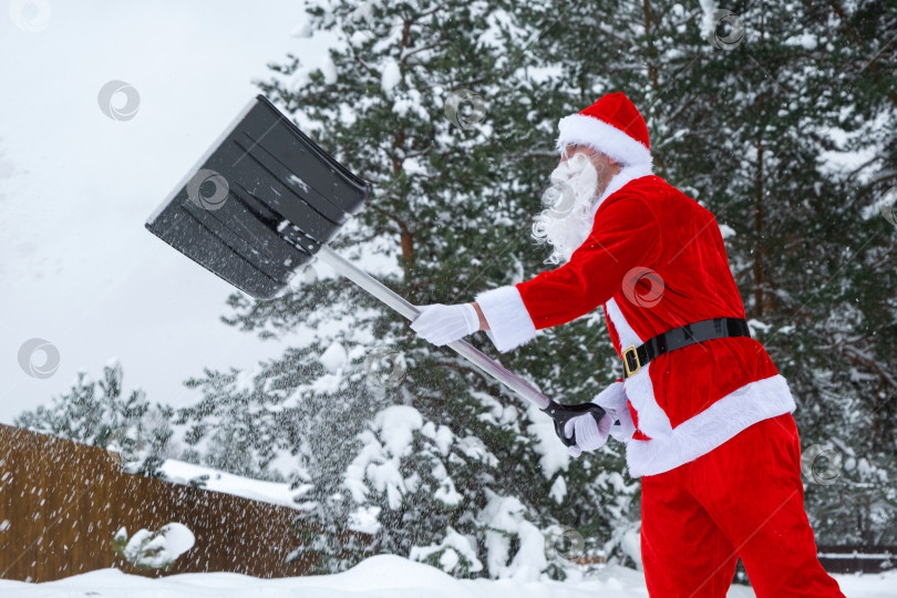 Скачать Санта Клаус убирает лопатой снег зимой на открытом воздухе после снегопада. Уборка улиц в деревне, расчистка проезда для автомобилей, сложные погодные условия на Рождество и Новый год фотосток Ozero