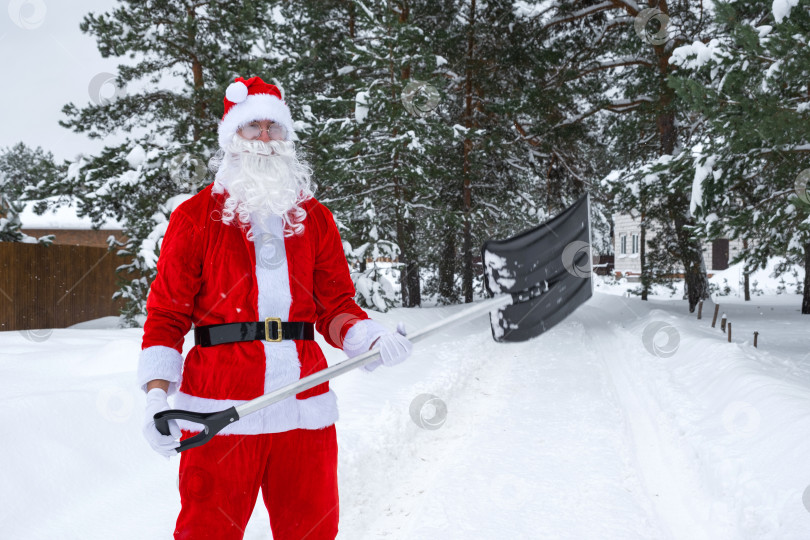 Скачать Санта Клаус убирает лопатой снег зимой на открытом воздухе после снегопада. Уборка улиц в деревне, расчистка проезда для автомобилей, сложные погодные условия на Рождество и Новый год фотосток Ozero