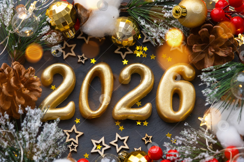 Скачать Золотые фигурки 2026, выполненные из свечей на фоне черного каменного сланца, украшены праздничным декором из звезд, блесток, еловых веток, шаров и гирлянд. Поздравительная открытка "с Новым годом". фотосток Ozero