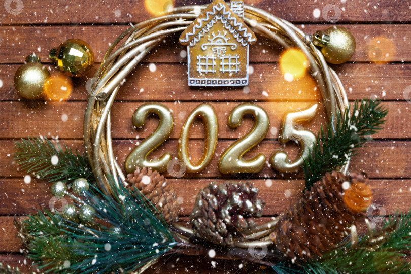 Скачать Новогодний ключ от дома с брелком коттедж на праздничном коричневом деревянном фоне с цифрой 2025 в венке, огни гирлянд. Покупка, строительство, переезд, ипотека, страхование фотосток Ozero