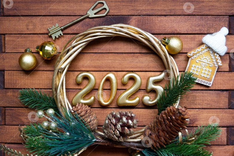 Скачать Новогодний ключ от дома с брелком коттедж на праздничном коричневом деревянном фоне с цифрой 2025 в венке, огни гирлянд. Покупка, строительство, переезд, ипотека, страхование фотосток Ozero