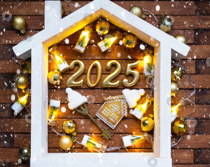 Скачать Ключ от дома с брелком коттедж на праздничном коричневом деревянном фоне со звездами, огоньками гирлянд. Новый 2025 год золотыми буквами под крышей. Покупка, строительство, переезд, ипотека, страхование фотосток Ozero