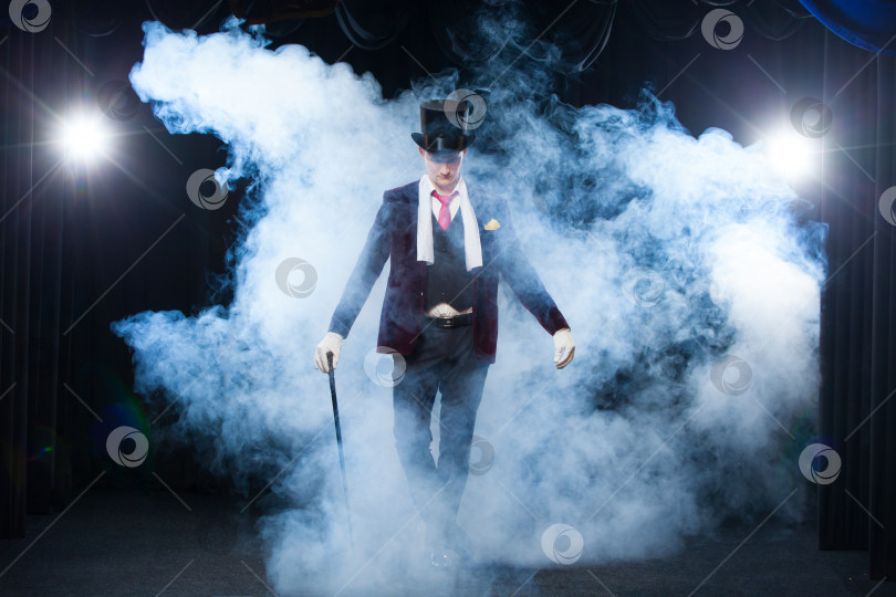Скачать Фокусник, Жонглер, Забавный человек, Черная магия, Иллюзия, стоящий на сцене с тростью из прекрасного света. окутанный прекрасным таинственным дымом фотосток Ozero