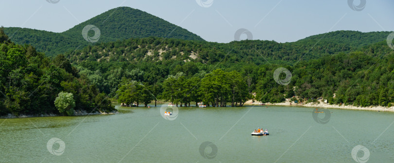 Скачать Живописный пейзаж с изумрудным кипарисовым озером и Кавказскими горами. Люди катаются на водных катамаранах и лодках. Популярное место отдыха недалеко от морского курорта Суко. Анапа, Россия - 20 июля 2021 года фотосток Ozero