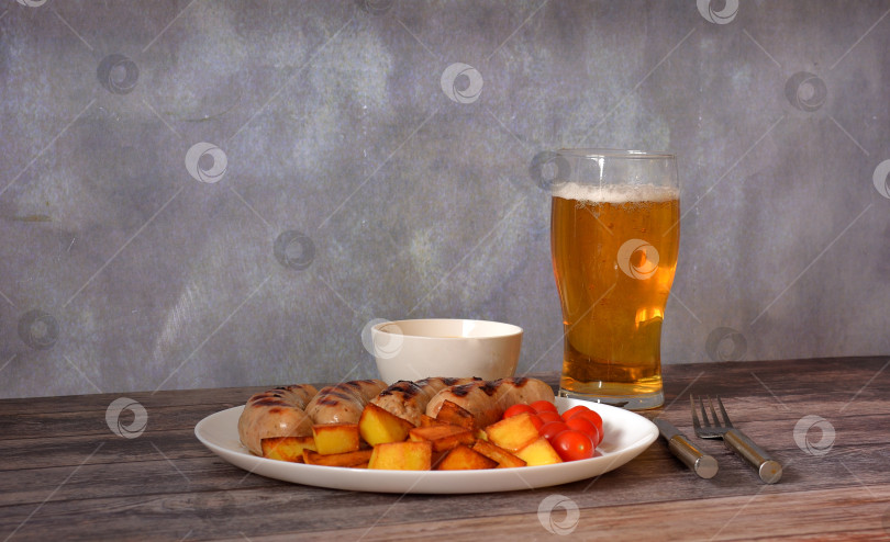 Скачать Бокал светлого пива и тарелка жареных сосисок-гриль с картофелем и помидорами на деревянном столе. фотосток Ozero