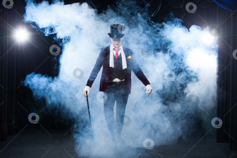 Скачать Фокусник, Жонглер, Забавный человек, Черная магия, Иллюзия, стоящий на сцене с тростью из прекрасного света. окутанный прекрасным таинственным дымом фотосток Ozero