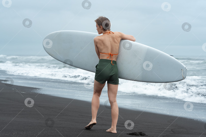 Скачать Вид сзади на женщину-серфингистку, идущую по песчаному пляжу с доской для серфинга в руках на фоне океанских волн фотосток Ozero