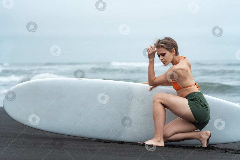 Скачать Женщина-серфер сидит перед доской для серфинга на пляже, глядя вниз в красивой сексуальной позе фотосток Ozero