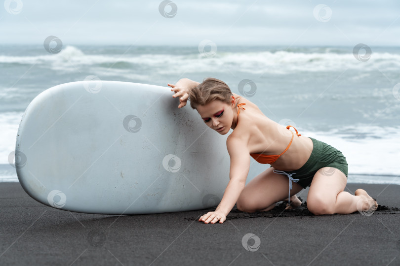 Скачать Женщина-серфер стоит на коленях на черном песке пляжа, держа доску для серфинга в расслабленной сексуальной позе фотосток Ozero