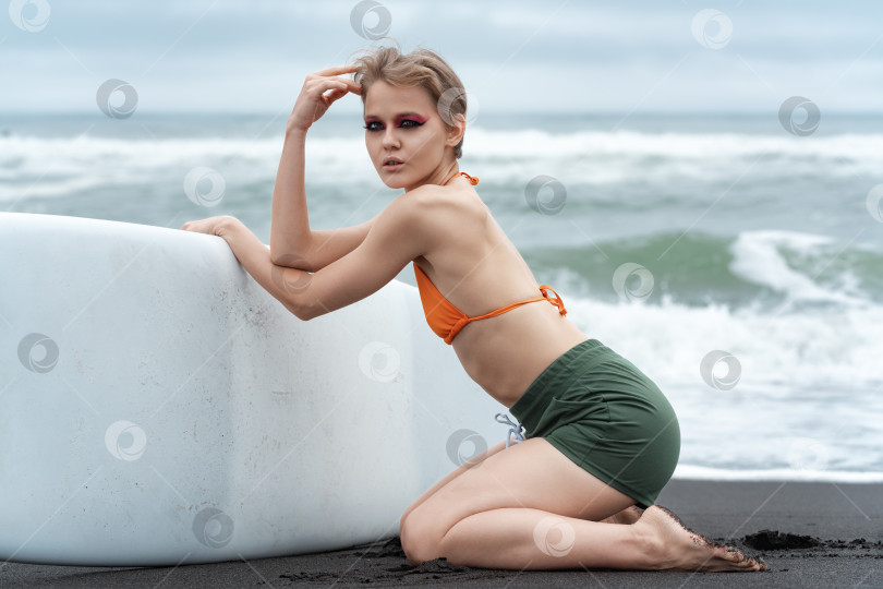 Скачать Стройная женщина-серфер выглядит потрясающе, когда она стоит на коленях на черном песке пляжа, держа в руках свою доску для серфинга фотосток Ozero