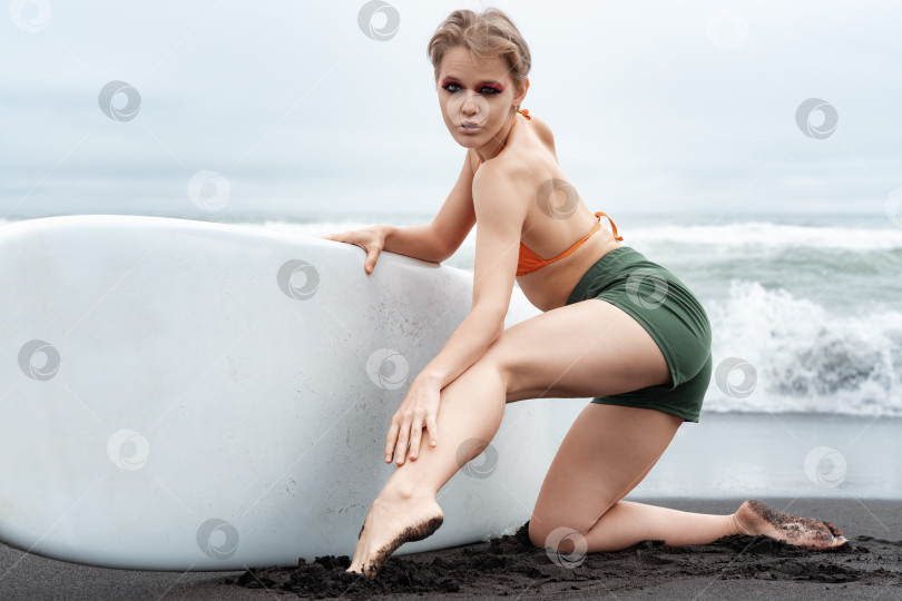 Скачать Женщина-серфер излучает уверенность, стоя на коленях на пляже с черным песком и легко держа доску для серфинга фотосток Ozero