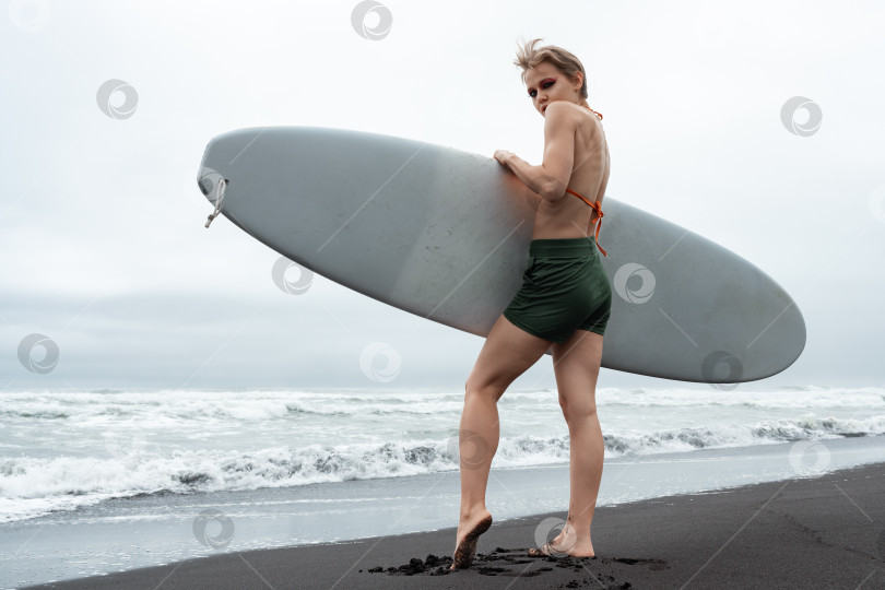 Скачать Женщина-серфер, стоящая на черном песчаном пляже с белой доской для серфинга в руках на фоне морских волн фотосток Ozero