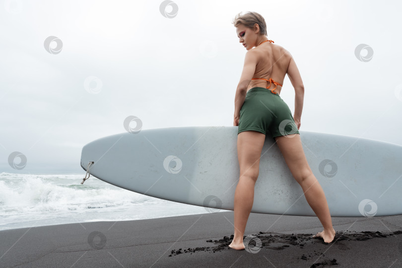 Скачать Вид сзади на женщину-серфингистку, стоящую на пляже, держащую доску для серфинга на фоне морских волн фотосток Ozero