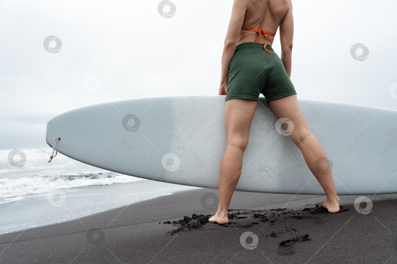 Скачать Женщина-серфер с доской для серфинга в руках, стоящая на черном песчаном пляже. Вид ног и ягодиц под низким углом фотосток Ozero