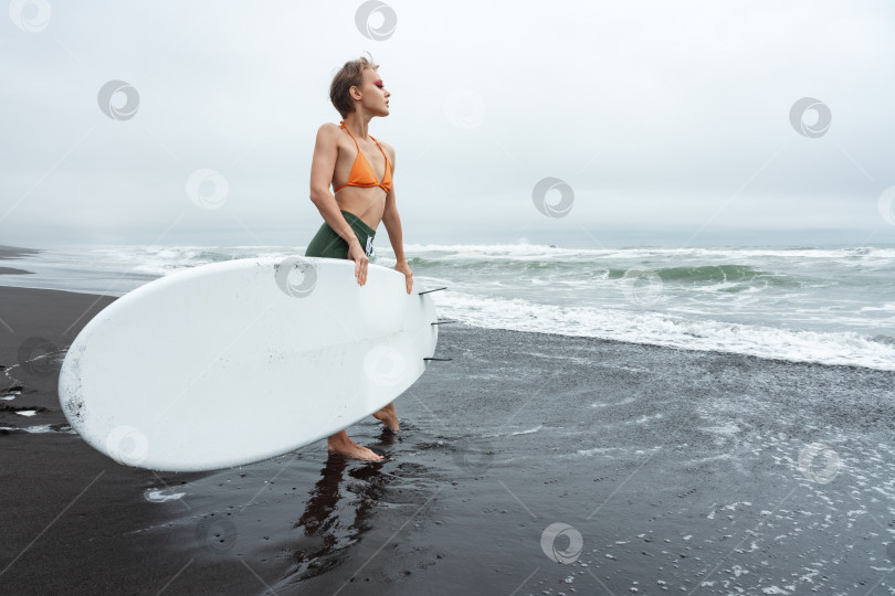 Скачать Серфер, стоящий на пляже с доской для серфинга в руках во время летних каникул. Стройная женщина смотрит на океанские волны фотосток Ozero