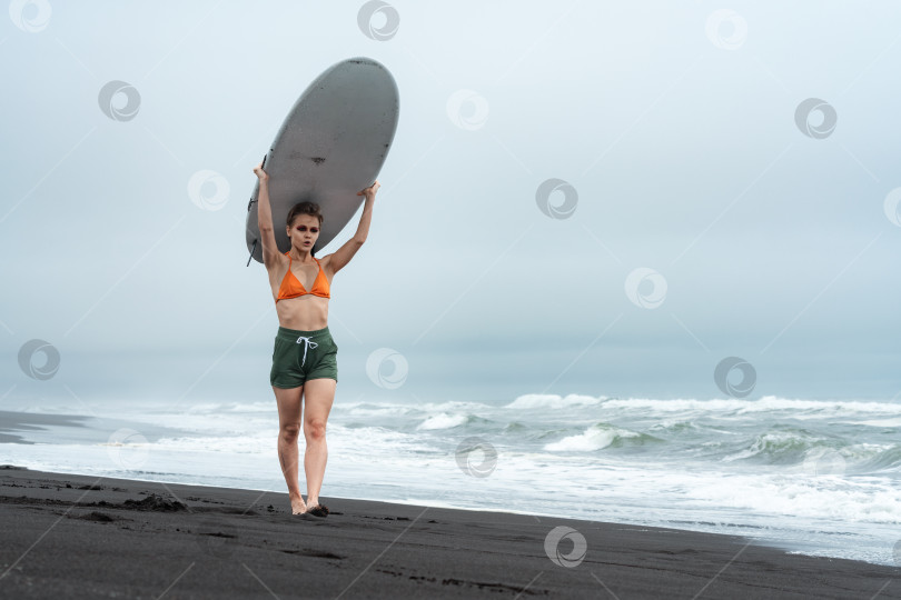 Скачать Женщина-серфер прогуливается по черному песчаному пляжу с белой доской для серфинга на голове во время летних каникул фотосток Ozero