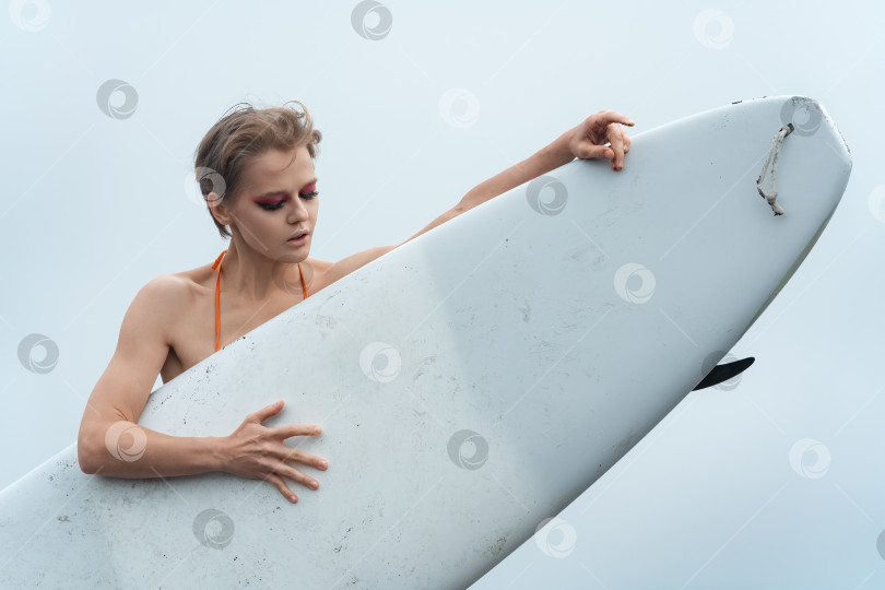 Скачать Женщина-спортивная фотомодель с доской для серфинга в руках. Она стоит за доской для серфинга и смотрит вниз фотосток Ozero