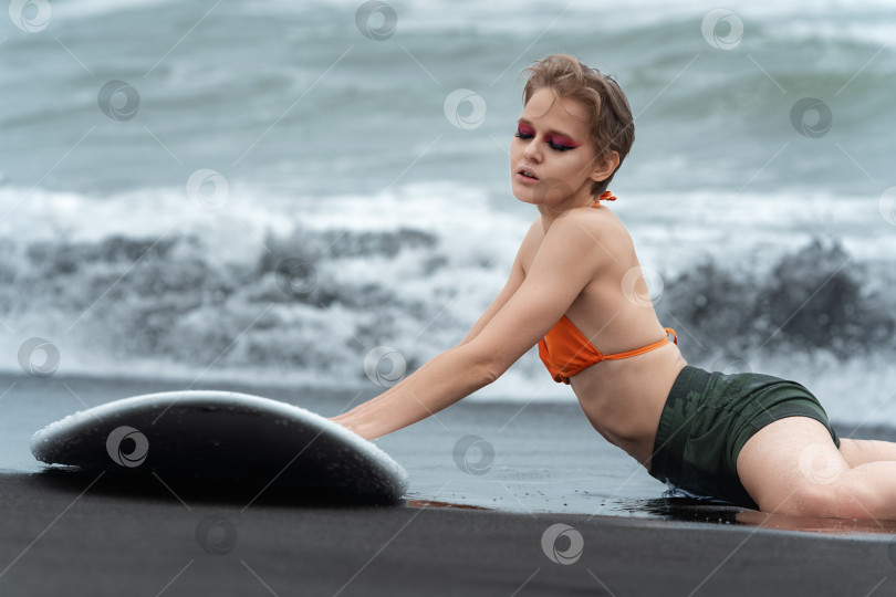 Скачать Очень горячая сексуальная женщина-серфер лежит на черном песке пляжа с доской для серфинга и смотрит вниз фотосток Ozero