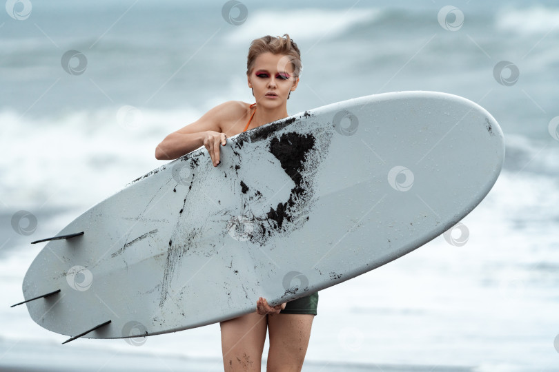 Скачать Женщина-серфер с белой доской для серфинга, идущая по пляжу, смотрит в камеру на фоне морских волн фотосток Ozero