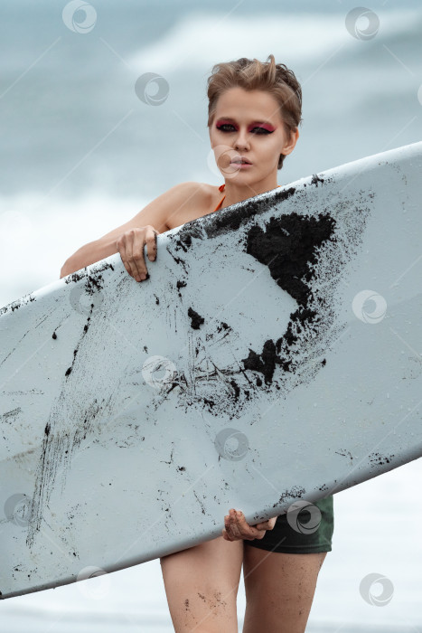 Скачать Женщина-серфер с доской для серфинга прогуливается по пляжу, глядя в камеру с морскими волнами на заднем плане фотосток Ozero
