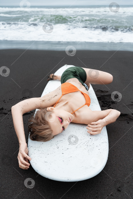 Скачать Женщина-серфер лежит на доске для серфинга на черном песчаном пляже. С закрытыми глазами и в расслабленной позе фотосток Ozero