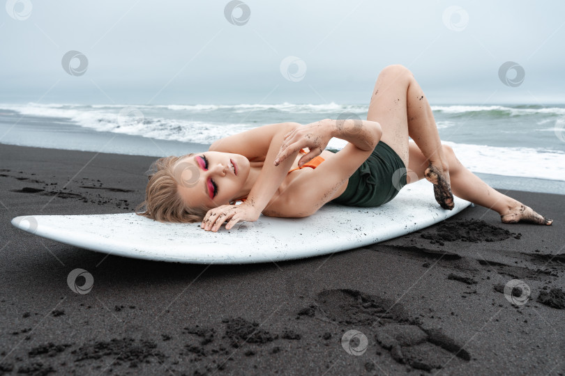 Скачать Женщина-серфер, лежащая на белой доске для серфинга на черном песчаном пляже. Спортивная фотомодель с закрытыми глазами фотосток Ozero