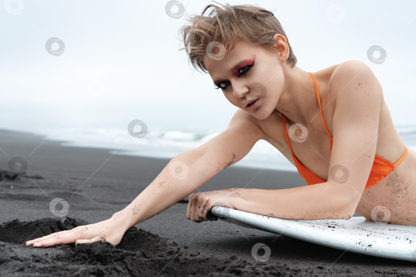 Скачать Портрет светловолосой женщины-серфингистки с ярким макияжем, лежащей перед доской для серфинга на песчаном пляже фотосток Ozero