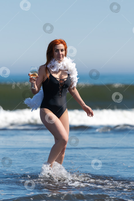 Скачать Рыжеволосая женщина в черном купальнике стоит в океанских волнах глубиной по щиколотку, держит бокал вина и улыбается фотосток Ozero