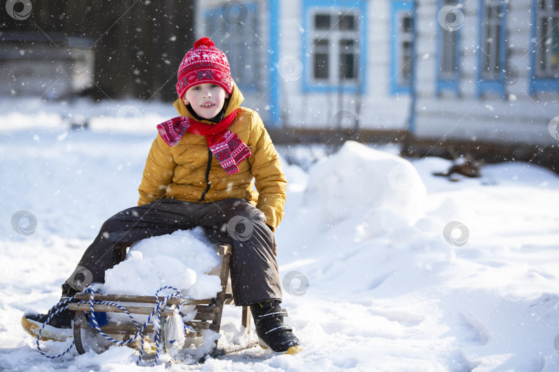 Скачать Маленький забавный мальчик зимним днем катается на деревянных санках по деревне. фотосток Ozero