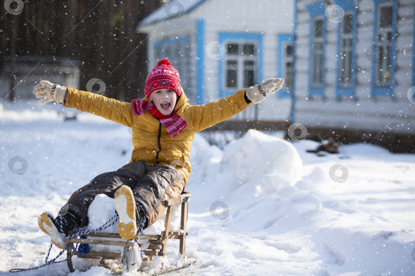 Скачать Маленький забавный мальчик зимним днем катается на деревянных санках по деревне. фотосток Ozero