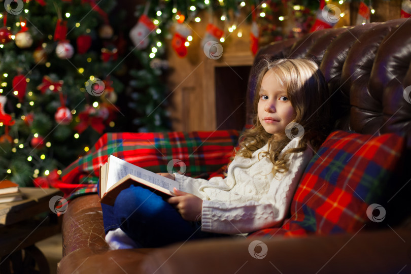 Скачать Счастливая маленькая девочка читает книгу рассказов на диване в уютной темной гостиной в канун Рождества, украшенная елка. Празднование Рождества дома. фотосток Ozero