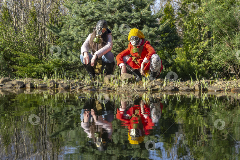 Скачать Маленькая девочка в красном со своей сестрой в газовом костюме сидит на корточках на берегу пруда. В зеркале пруда отражение девочек. фотосток Ozero