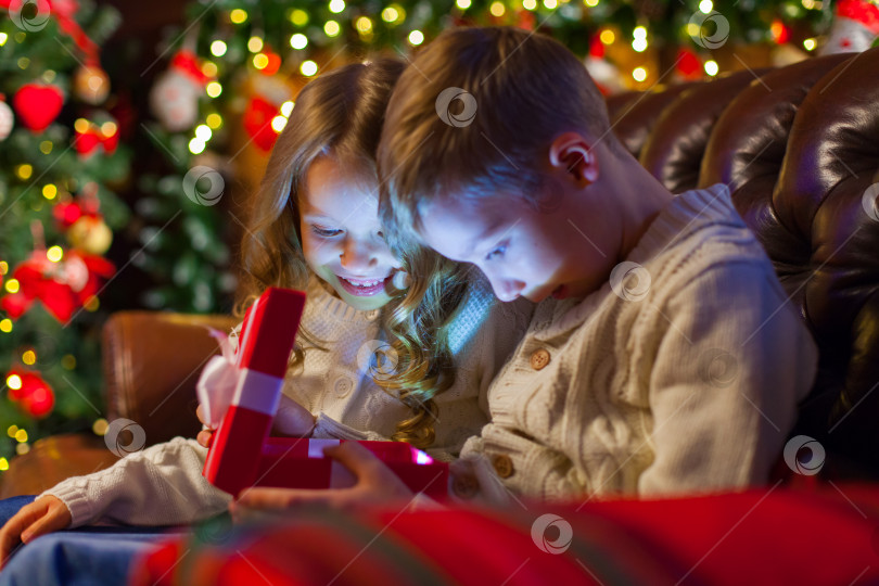 Скачать Рождественская волшебная подарочная коробка для детей. на диване в уютной темной гостиной в канун Рождества наряжается елка. Празднование Рождества дома. фотосток Ozero